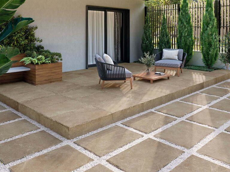 baldosa rústica para exterior pulse antique noce instalada en una terraza que combina una zona de plataforma elevada con muebles de exterior y un camino de baldosas colocadas sobre grava blanca, rodeado de vegetación y cipreses