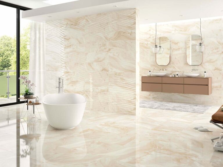 baño efecto mármol brillante diva cream con paredes y suelo coordinados, una bañera exenta blanca y un mueble de lavabo de madera, destacando una zona de pared con azulejos decorativos de relieve lineal que aportan textura al diseño