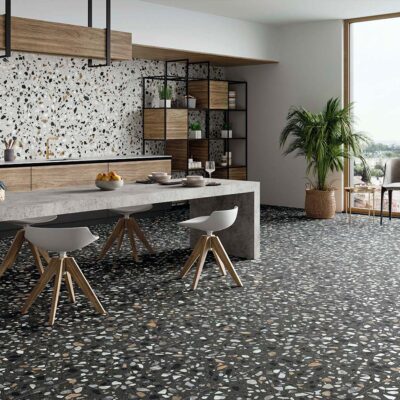 Azulejo de pared Crisp XL White, con efecto terrazo sobre base blanca y fragmentos multicolores (negro, gris, ocre), instalado como revestimiento en la pared de una moderna cocina-comedor, contrastando con el suelo de terrazo oscuro.