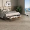 Dormitorio de interior con suelo y pared de cabecero revestidos con baldosa Caliope Beige, efecto piedra en tono beige claro con sutiles inclusiones, y acceso a balcón con vistas al mar.