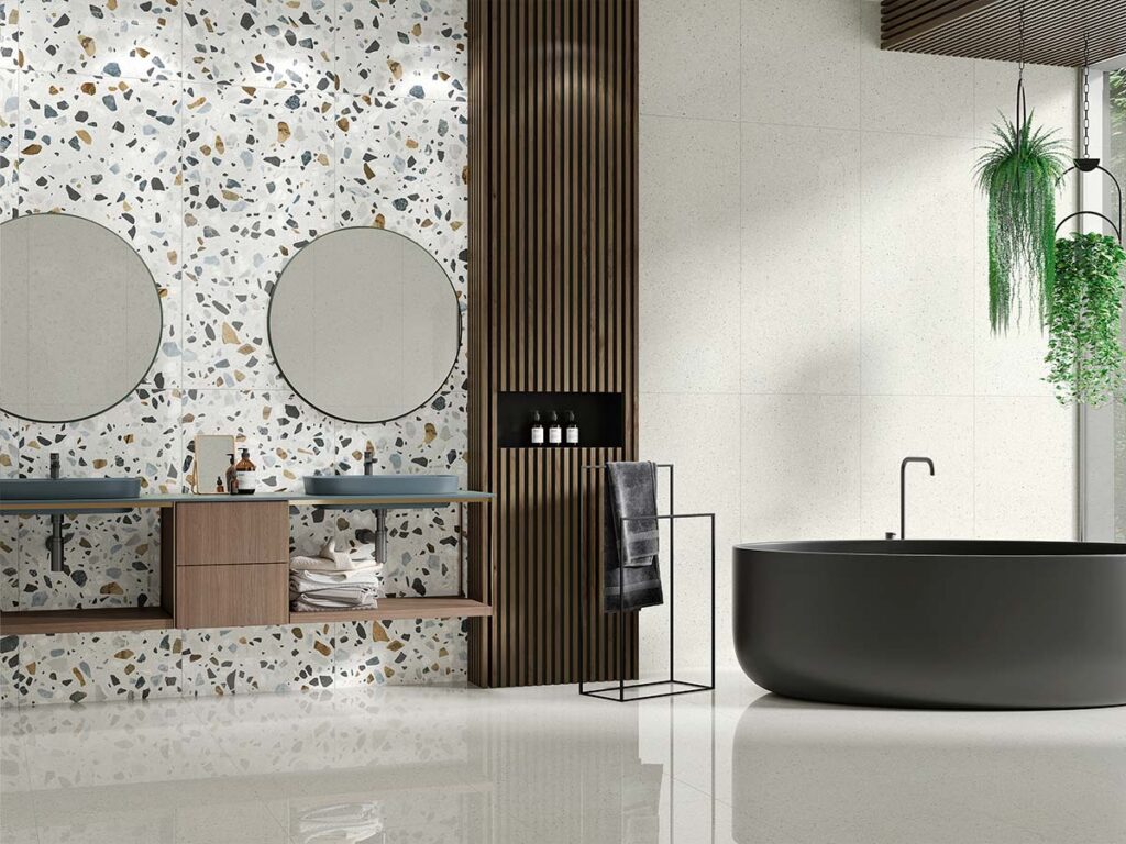 Azulejo de pared Crisp XL White, con efecto terrazo sobre base blanca y fragmentos multicolores (negro, gris, ocre), instalado como revestimiento en la pared de una moderna cocina-comedor, contrastando con el suelo de terrazo oscuro.