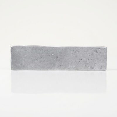 Azulejo rectangular gris porcelánico con textura de piedra sobre superficie reflectante.