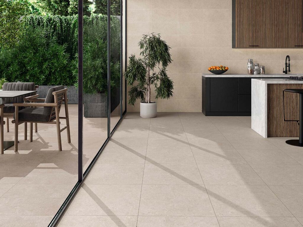 baldosa cerámica beige ulisse natural instalada en una cocina de diseño moderno con isla, que presenta un pavimento y paredes coordinados en tonos arena, muebles de cocina en negro mate y madera, y una conexión fluida hacia un comedor exterior rodeado de vegetación a través de una gran cristalera