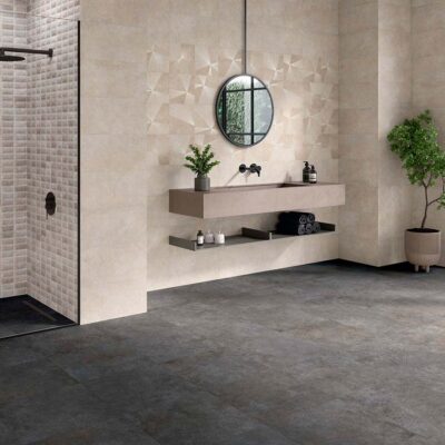 Paredes de un baño moderno revestidas con baldosas Feroe Greige imitación piedra, que combinan un acabado liso, un decorado geométrico y un mosaico en la ducha, en contraste con un suelo oscuro.