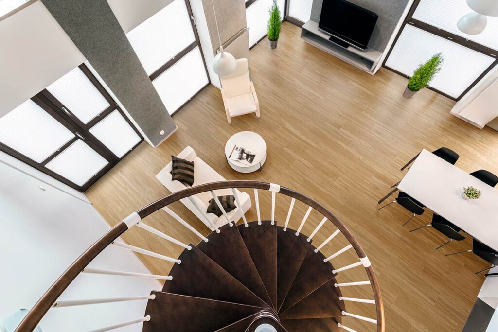 Vista aérea de un salón moderno con suelo porcelánico imitación madera rectificado leroy merlin, una escalera de caracol y grandes ventanales.