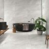 Baño moderno con suelo y paredes revestidos en porcelánico Sakhir Grey, un elegante efecto piedra en tono gris claro con sutiles vetas blancas, contrastando con lavabos y bañera oscuros.