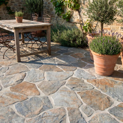Un espacio exterior de estilo rústico con suelo de piedra natural de tonos grises y marrones, formato irregular y acabado mate, con juntas blancas.