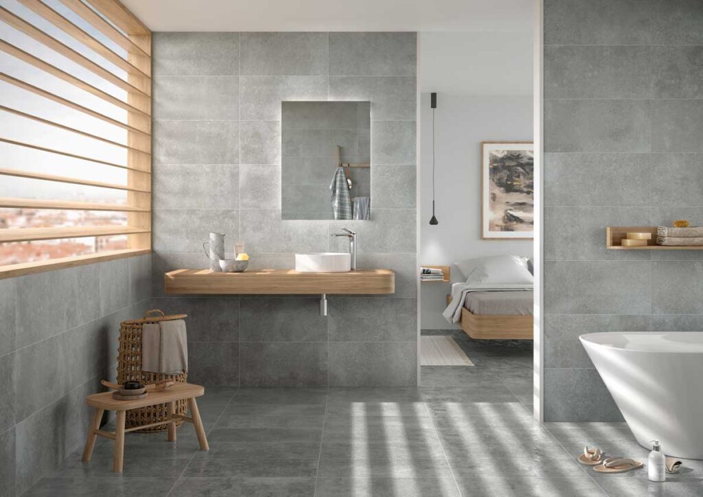 Baño moderno con suelo y paredes de suelo porcelanico efecto cemento claro. Mueble de madera, lavabo y bañera.