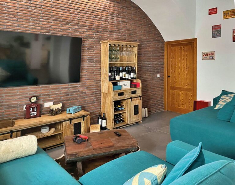 Salón con pared de revestimiento de ladrillo rojo visto, suelos porcelánicos de tonalidad oscura, mueble bar de madera con botellas, sofá turquesa y televisión empotrada en pared.