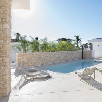 Terraza exterior con suelo porcelánico en tonos claros, piscina rectangular, tumbonas blancas y revestimiento de hormigón imitación piedra en tonos beige. Zona de estar con moderna edificación blanca y vegetación tropical de fondo.