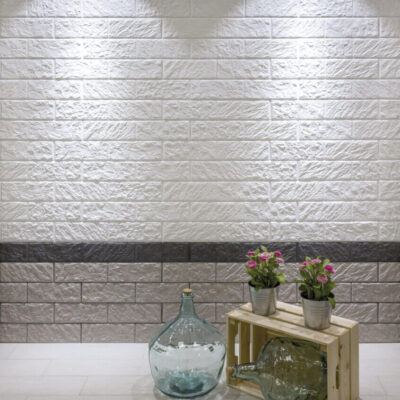 Cocina contemporánea con revestimiento de pared en tonos grises y blancos con textura efecto ladrillo. Franja decorativa en gris oscuro separa dos áreas de diferentes tonalidades, con suelo blanco y accesorios de madera natural con plantas decorativas en primer plano.