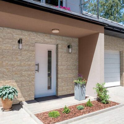 Entrada de vivienda moderna con revestimiento de pared efecto piedra en tonos marfil y beige, puerta de acceso blanca con vidrio, maceteros decorativos y pavimento de hormigón en tonos terracota.