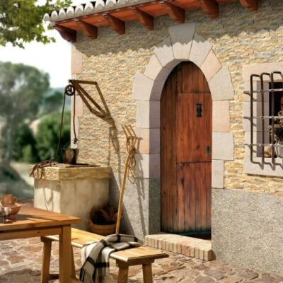 Espacio exterior con aplacado de piedra natural en tonos beige y gris formando una fachada rústica, puerta de madera oscura con arco de dovelas, ventana con reja de hierro y mesa de madera con elementos decorativos tradicionales.