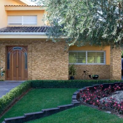 Vivienda unifamiliar contemporánea con fachada de aplacado de ladrillo cerámico visto en tonos marrones, puerta de entrada arqueada, ventanales y jardín con flores rojas y árbol centenario.