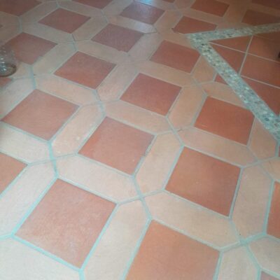 Pavimento interior con baldosas de barro cocido en formato rombo y cuadrado, tonos arena y rojo terracota con acabado rústico natural, dispuestas en patrón geométrico alternado.