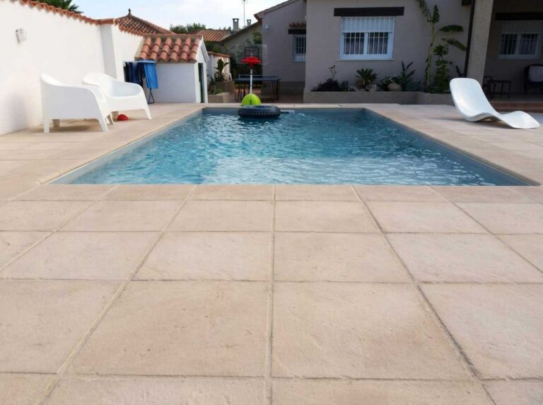 Terraza exterior con baldosas de color ámbar cálido junto a una piscina rectangular. El suelo presenta un acabado rústico y tonos tierra, ideal para áreas de piscina y jardín, con tumbonas blancas y vegetación tropical de fondo.