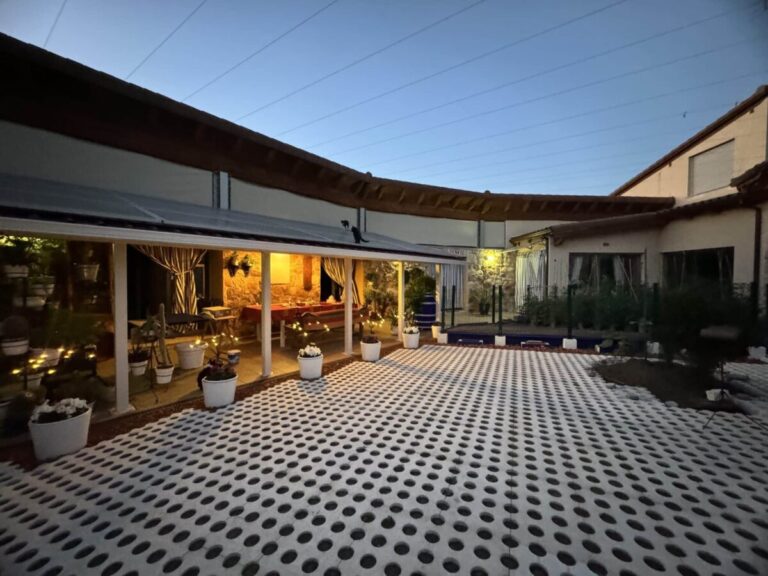 Terraza exterior con pavimento de celosía de hormigón en tonos grises y blancos, cubierta por pérgola de madera. A la izquierda, zona de estar iluminada con mobiliario de interior visible a través de cristaleras, y macetas blancas decorativas distribuidas en el suelo.