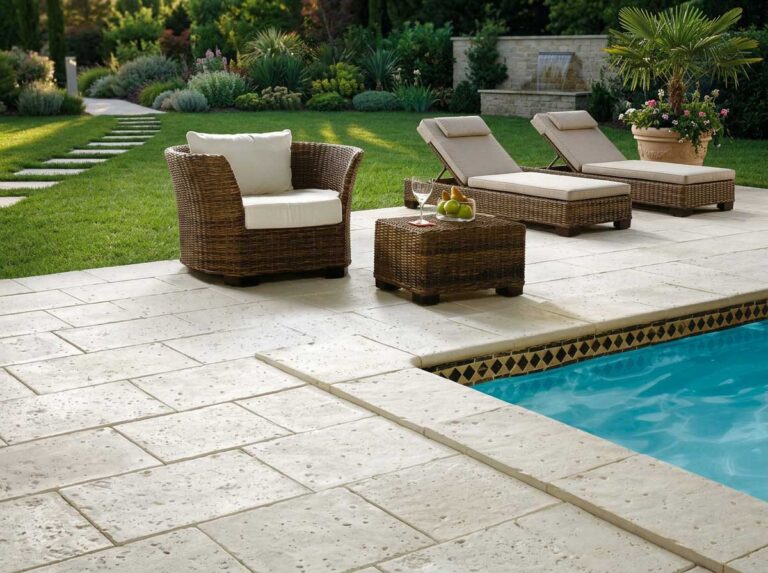 Terraza junto a piscina con pavimento porcelánico en tonos claros, mobiliario de rattán con cojines beige y tumbonas de exterior. Al fondo, jardín con palmeras y aplacado de hormigón decorativo.