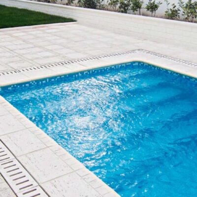 Terraza exterior junto a piscina con pavimento de grandes losas cerámicas en tonos crudo y beige. Franja decorativa de rejilla metálica gris bordeando el perímetro de la piscina. Césped artificial visible en el fondo y plantas en macetas.