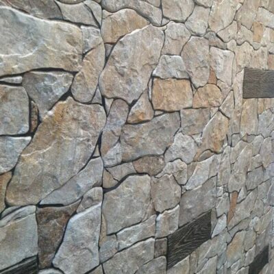 Superficie de gres cerámico con acabado mate imitando piedra natural, tonalidades beige y grises con veteado irregular y textura rugosa que simula mampostería rústica.