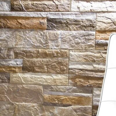 Pared revestida con gres porcelánico efecto piedra natural en tonos beige y dorados, con textura rugosa y formato rectangular en colocación de soga. Detalle de silla tapizada blanca en primer plano.
