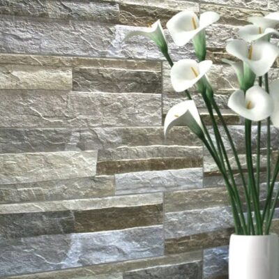 Pared revestida con aplacado efecto piedra natural en tonos grises y beige, con un jarrón blanco de flores de lirio blancas en primer plano. El acabado texturizado simula piedra natural en formato de hiladas irregulares, ideal para suelo porcelánico de acabado similar.