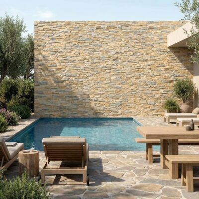 Terraza exterior con pared de revestimiento cerámico efecto piedra natural en tonos beige y grises, acompañada de piscina, mobiliario de madera y vegetación mediterránea.