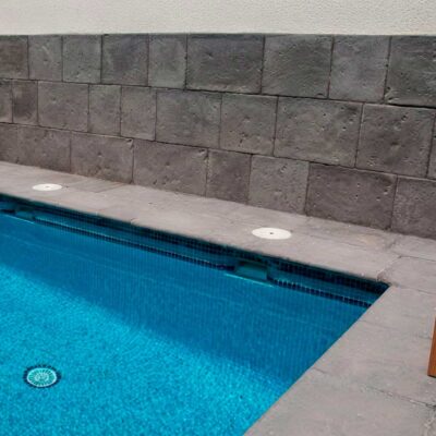 Terraza junto a piscina con revestimiento de pared en aplacado de hormigón color antracita texturizado. Piscina con agua turquesa, toallas blancas y plantas suculentas sobre decking de hormigón.