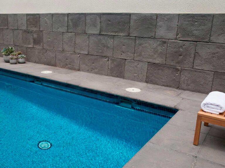 Terraza junto a piscina con revestimiento de pared en aplacado de hormigón color antracita texturizado. Piscina con agua turquesa, toallas blancas y plantas suculentas sobre decking de hormigón.