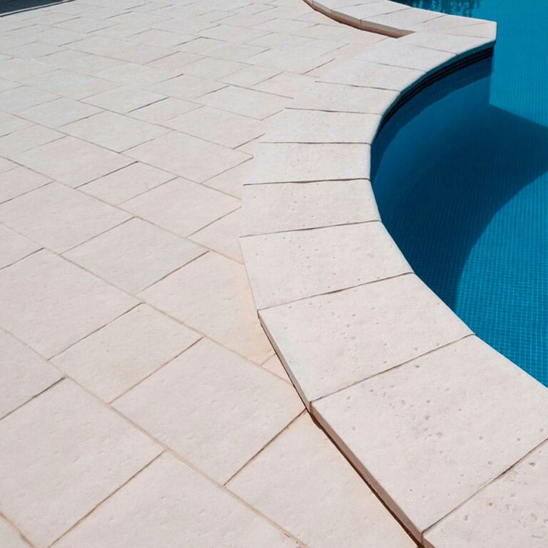 Terraza junto a piscina con revestimiento de baldosa efecto mármol en tonos crema y beige, dispuesta en patrón diagonal. El pavimento rodea el borde curvo de la piscina con agua azul turquesa.