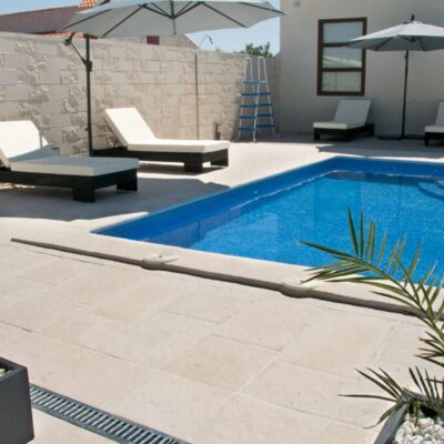 Terraza exterior junto a piscina con pavimento de hormigón en tonos grises. Se aprecian tumbonas con sombrilla, revestimiento de pared con textura en tonalidad beige y detalles de paisajismo con plantas ornamentales.