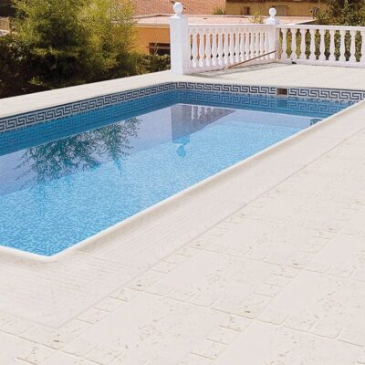Piscina rectangular con agua azul clara, bordeada por pavimento de cerámica beige claro en formato cuadrado, rematada con cenefa decorativa de patrón griego en azul y blanco. Balconada blanca y vegetación al fondo.
