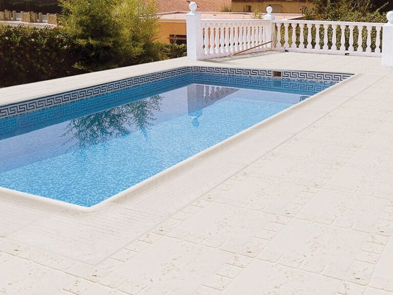 Piscina rectangular con agua azul clara, bordeada por pavimento de cerámica beige claro en formato cuadrado, rematada con cenefa decorativa de patrón griego en azul y blanco. Balconada blanca y vegetación al fondo.