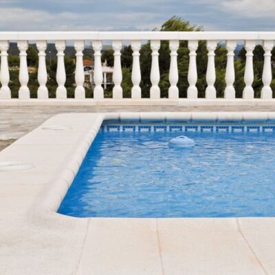 Terraza junto a piscina rectangular con pavimento porcelánico de gran formato en tonos beige claros. Balustrada blanca de balaustres clásicos y vistas al paisaje con vegetación de cipreses al fondo.