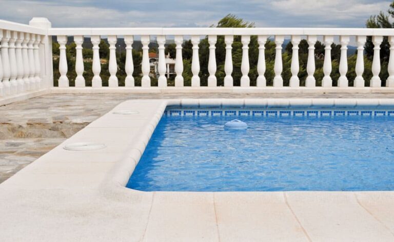 Terraza junto a piscina rectangular con pavimento porcelánico de gran formato en tonos beige claros. Balustrada blanca de balaustres clásicos y vistas al paisaje con vegetación de cipreses al fondo.