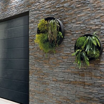 Fachada exterior con aplacado Revestimiento Andorra Marengo en tonos grises y marrones, textura de piedra natural, junto a puerta metálica negra y macetas colgantes con plantas verdes.
