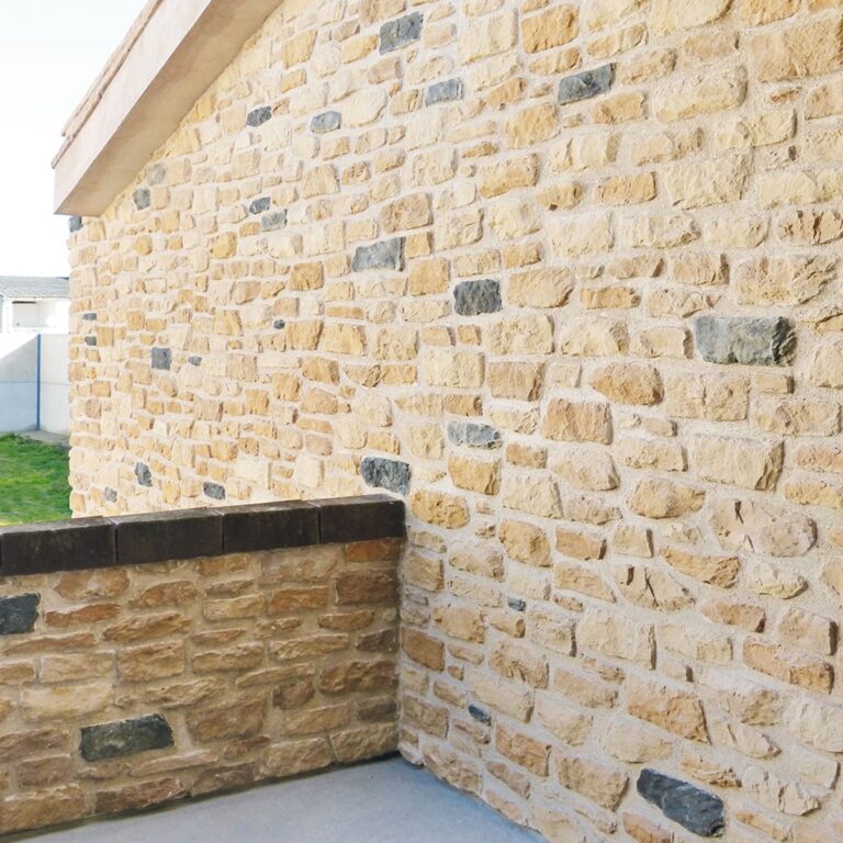 Esquina exterior con revestimiento de pared en tonos beige y gris, mostrando un efecto ladrillo con piezas rectangulares irregulares y acabado rústico natural. Incluye zócalo de protección oscuro en la base y vista parcial de estructura blanca y zona de césped.