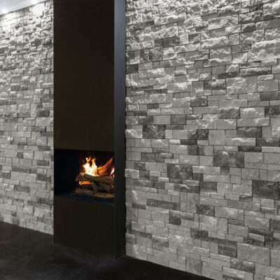 Espacio interior con paredes revestidas de aplacado gris efecto piedra natural en formato rectangular, iluminado con apliques empotrados. Chimenea de pared con fuego visible integrada en el revestimiento, creando un contraste dramático entre la textura de piedra y la llama.