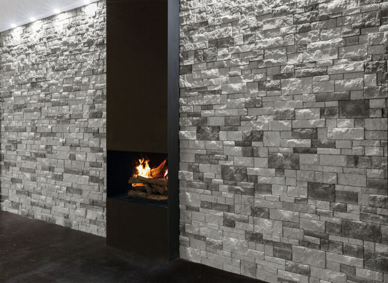 Espacio interior con paredes revestidas de aplacado gris efecto piedra natural en formato rectangular, iluminado con apliques empotrados. Chimenea de pared con fuego visible integrada en el revestimiento, creando un contraste dramático entre la textura de piedra y la llama.