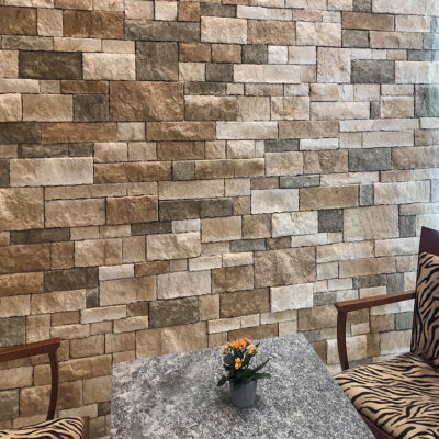 Espacio interior con pared de aplacado de hormigón en tonos beige, marrón y gris con textura de piedra natural. Mobiliario de madera con tapicería de rayas cebra y mesa de granito gris con maceta de flores naranjas.