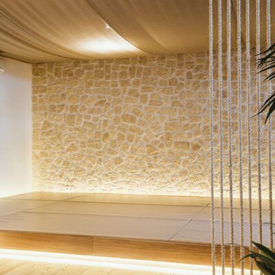 Interior contemporáneo con pared de piedra natural en tonos beige y suelo de gres porcelánico imitación madera. Divisor de cuerda blanca y iluminación integrada crean una atmósfera minimalista y elegante.
