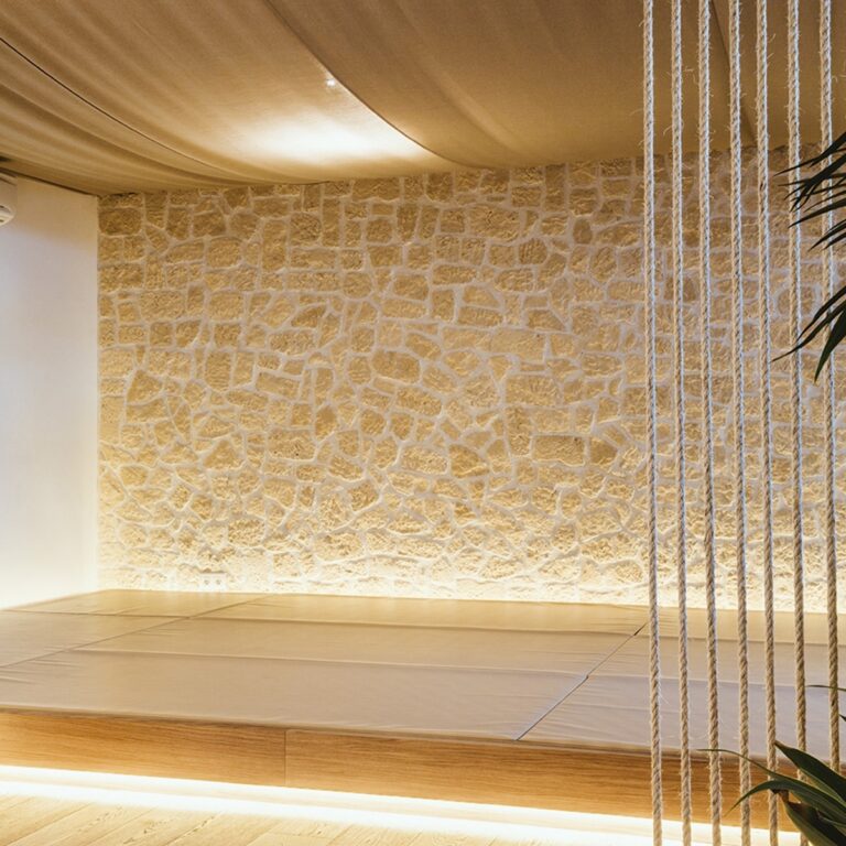 Interior contemporáneo con pared de piedra natural en tonos beige y suelo de gres porcelánico imitación madera. Divisor de cuerda blanca y iluminación integrada crean una atmósfera minimalista y elegante.