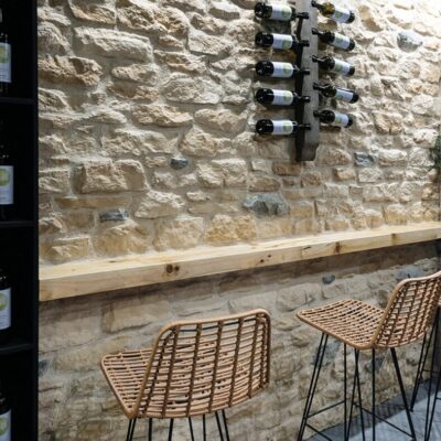 Bodega con pared de revestimiento interior imitación piedra en tonos beige y crema, con barriles de vino montados en estructura metálica. Dos taburetes de ratán con patas de metal frente al mostrador de madera iluminado con focos LED.