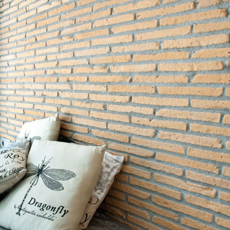 Pared interior revestida con ladrillo cerámico Cambridge Amanecer en tonos cálidos melocotón y beige, con textura rústica y juntas de mortero gris. Cojines decorativos con estampado de libélula reposan contra el revestimiento.