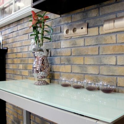 Cocina contemporánea con revestimiento de ladrillo cerámico Oxford Bristol en tonos ocre y marrón instalado en la pared trasera, complementado con encimera de vidrio blanco y accesorios decorativos.