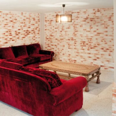 Salón interior con paredes revestidas de ladrillo cerámico visto en tonos claros y blancos, mobiliario de terciopelo rojo oscuro y banco de fibra natural. El revestimiento de ladrillo presenta un patrón de colocación horizontal que aporta calidez y textura al espacio.