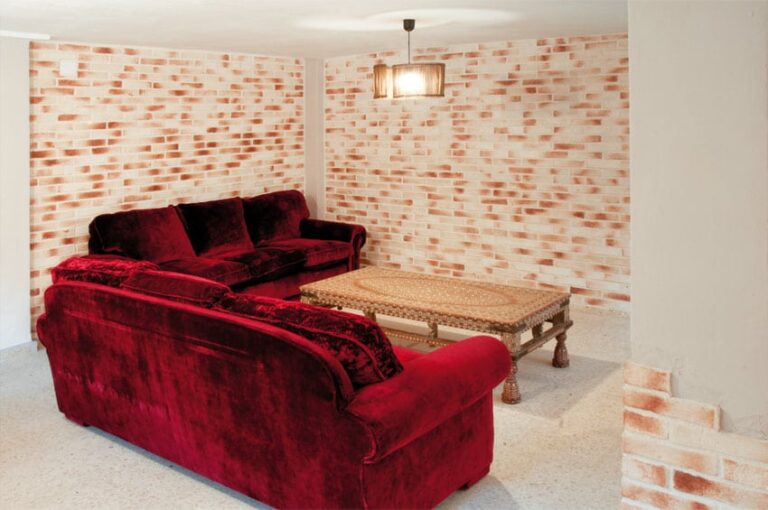 Salón interior con paredes revestidas de ladrillo cerámico visto en tonos claros y blancos, mobiliario de terciopelo rojo oscuro y banco de fibra natural. El revestimiento de ladrillo presenta un patrón de colocación horizontal que aporta calidez y textura al espacio.