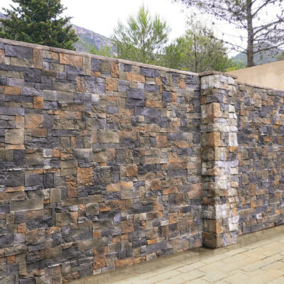 Aplacado de pared exterior con efecto piedra natural en tonos grises, marrones y dorados. El revestimiento Montblanc Otoño presenta un patrón de mampostería irregular con bloques de diferentes tamaños y acabado rústico texturizado, instalado en una zona ajardinada con árboles de fondo.