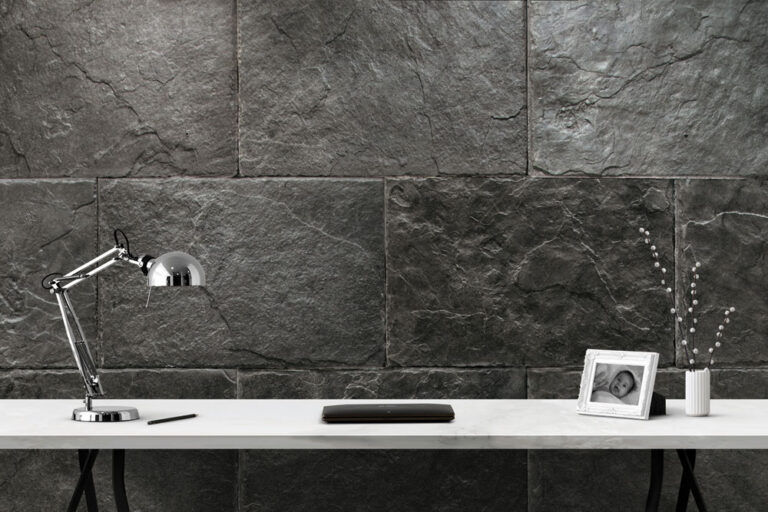Muro revestido con baldosas negras de efecto pizarra en formato rectangular, instaladas en patrón de tizon. La superficie muestra textura natural irregular y acabado mate, creando un fondo arquitectónico oscuro y minimalista junto a accesorios de escritorio.