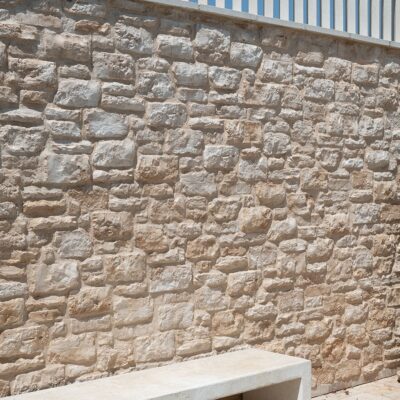 Superficie de revestimiento con textura de piedra natural irregular, tonos beige y gris claro, acabado rústico mate con relieve pronunciado que imita mampostería de cantera.